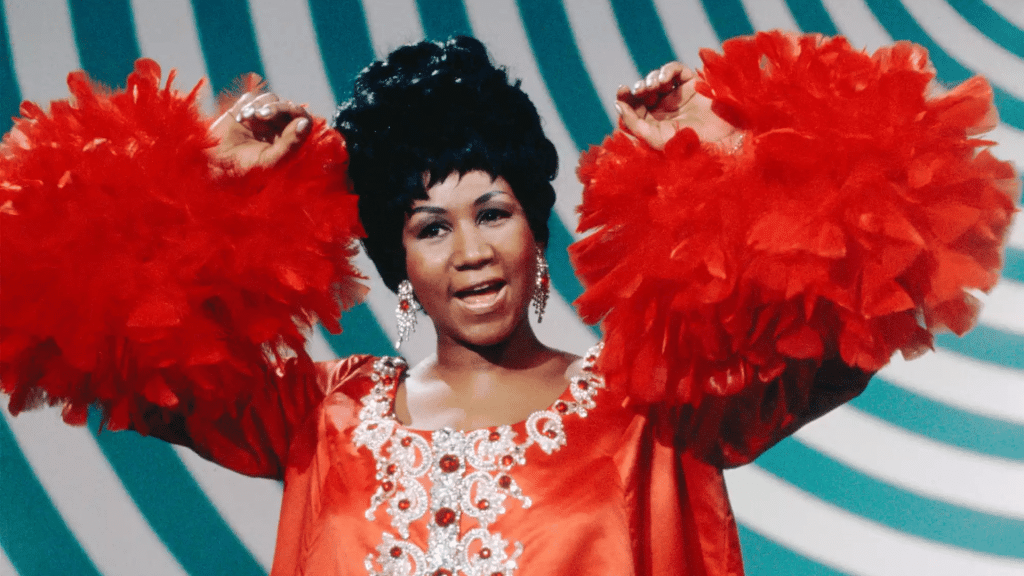 Aretha Franklin performing in a red outfit with feathered sleeves on stage.