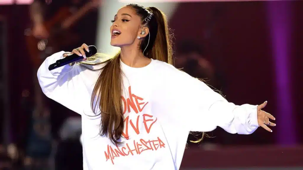Ariana Grande performing at the One Love Manchester benefit concert wearing a white sweatshirt on stage.