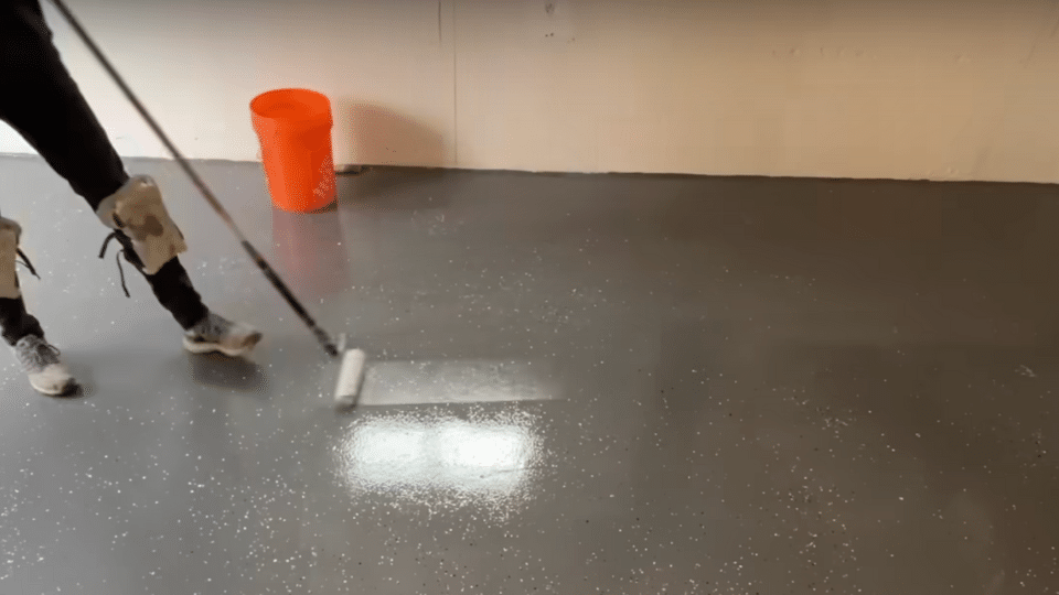 A person using a paint roller attached to an extension pole to apply a clear glossy topcoat over a grey epoxy garage floor.