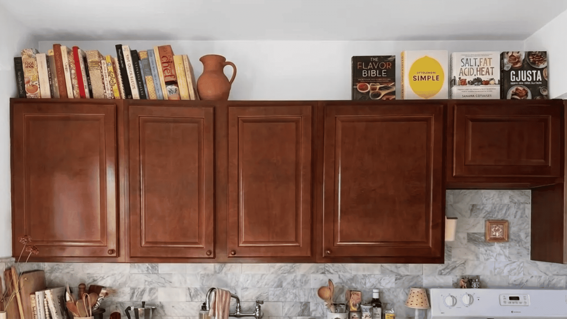 Cookbooks stacked and standing on top of dark wood kitchen cabinets beside a small ceramic pitcher