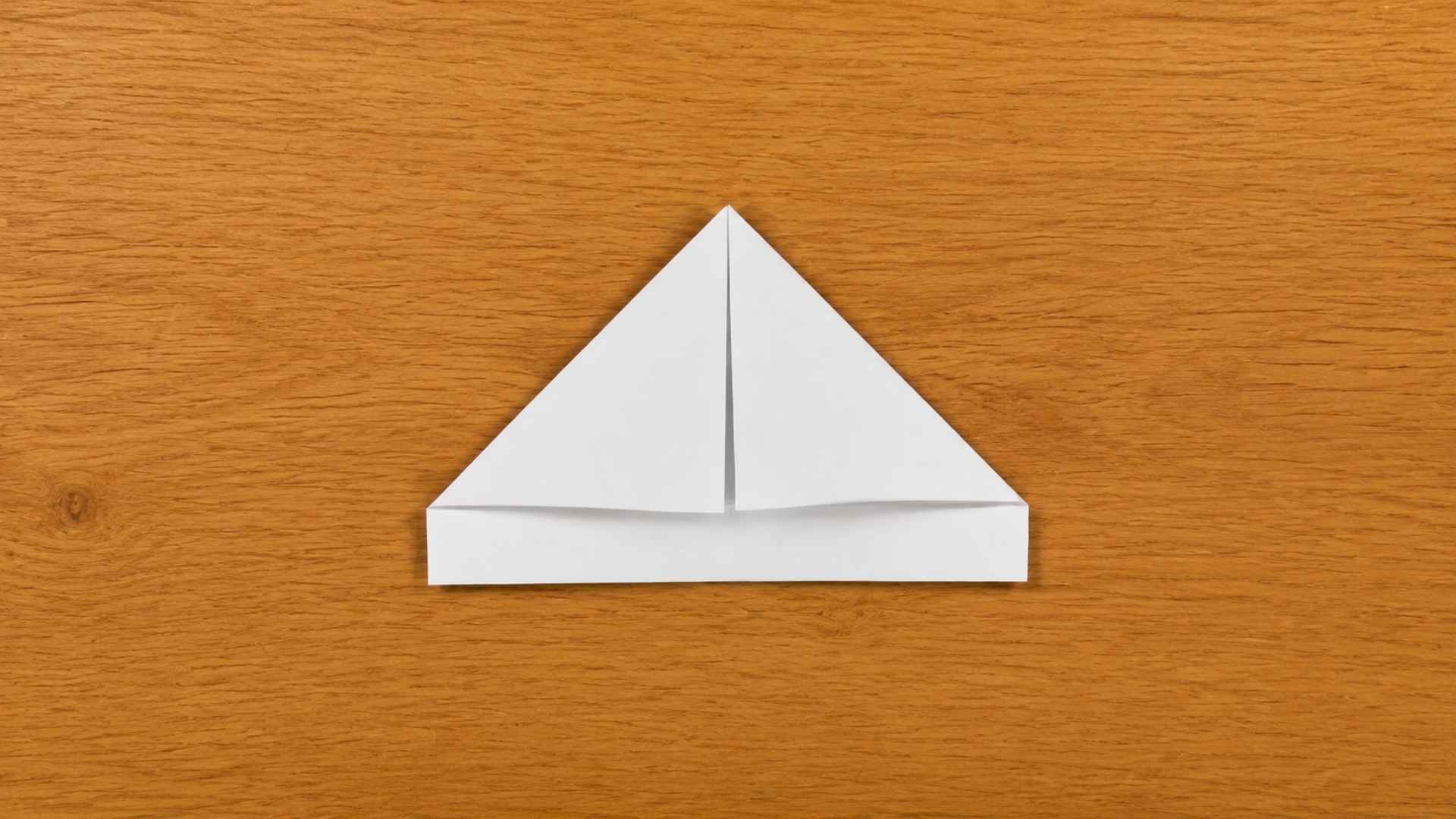 White paper folded into a triangular base shape (origami step 2) placed on a wooden table with warm natural lighting, showing a clean paper crease and symmetrical triangle form.