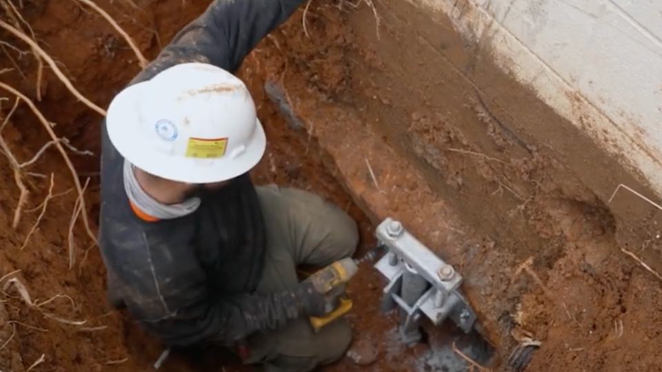 A worker wearing a white hard hat kneeling in an excavated dirt pit, using a tool to adjust a steel pier bracket attached to a concrete foundation.