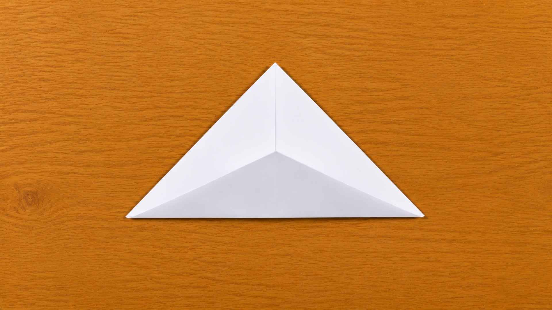 White origami paper folded into a triangular shape on a wooden table with soft natural lighting and subtle shadows.