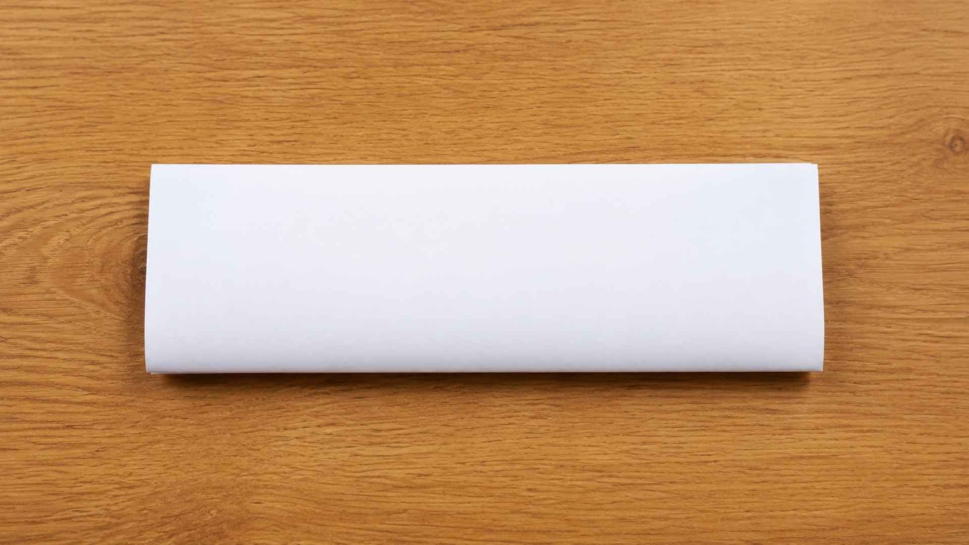 White paper folded into a neat rectangle on a wooden table showing Step 1 of making a paper boat origami tutorial.