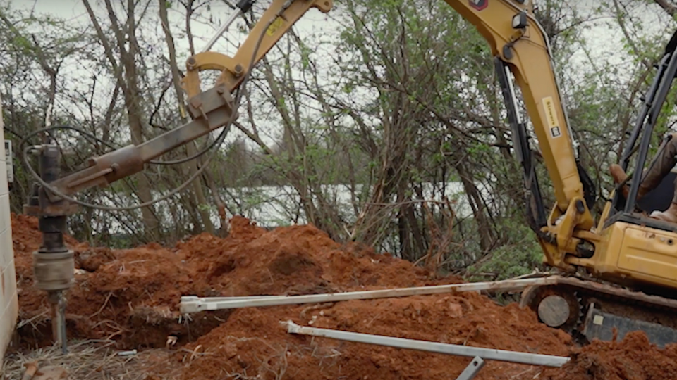 A yellow excavator with a specialized hydraulic pier-driving attachment working in an excavated dirt trench near trees at a construction site.