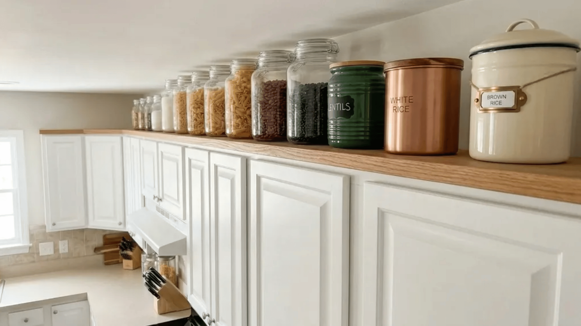 Glass jars and labeled canisters filled with dry goods lined up on top of white kitchen cabinets.