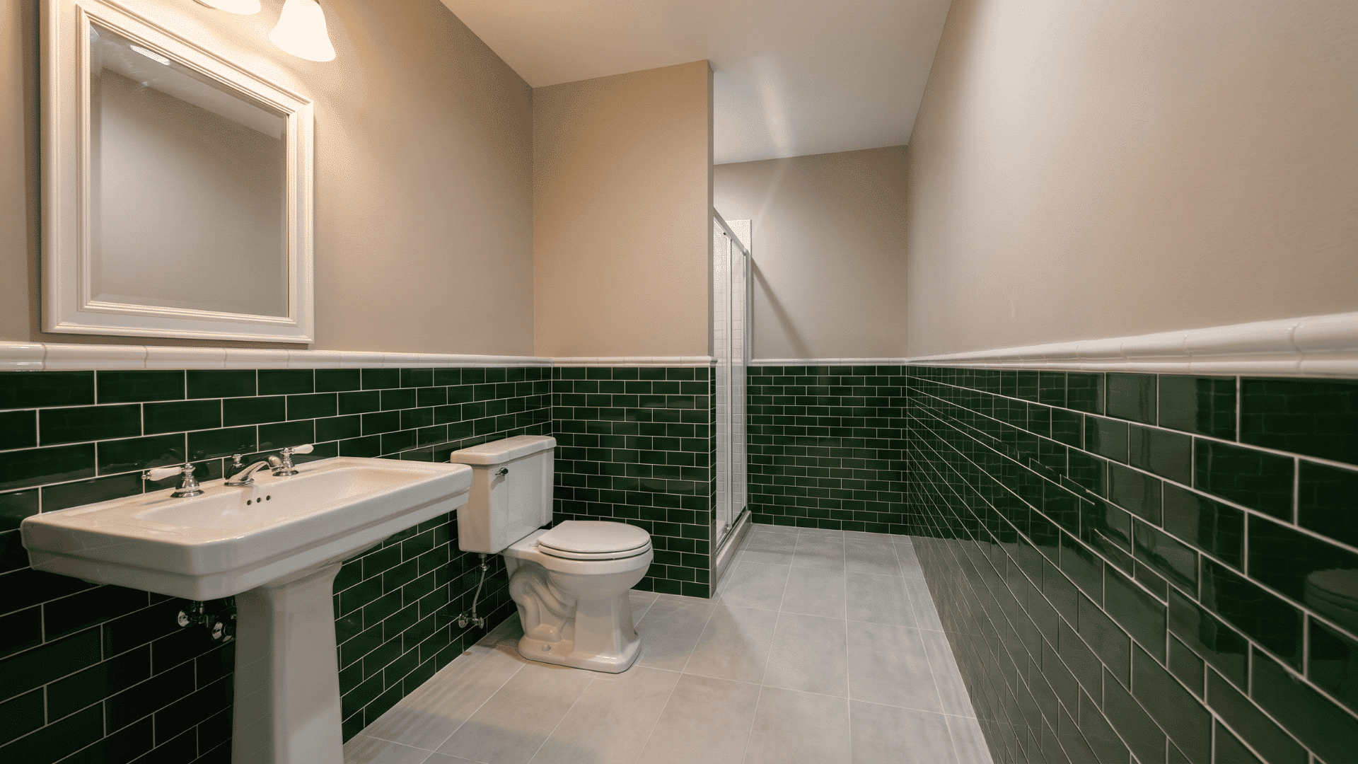 Bathroom with dark green tiled walls, pedestal sink, toilet, and framed mirror.