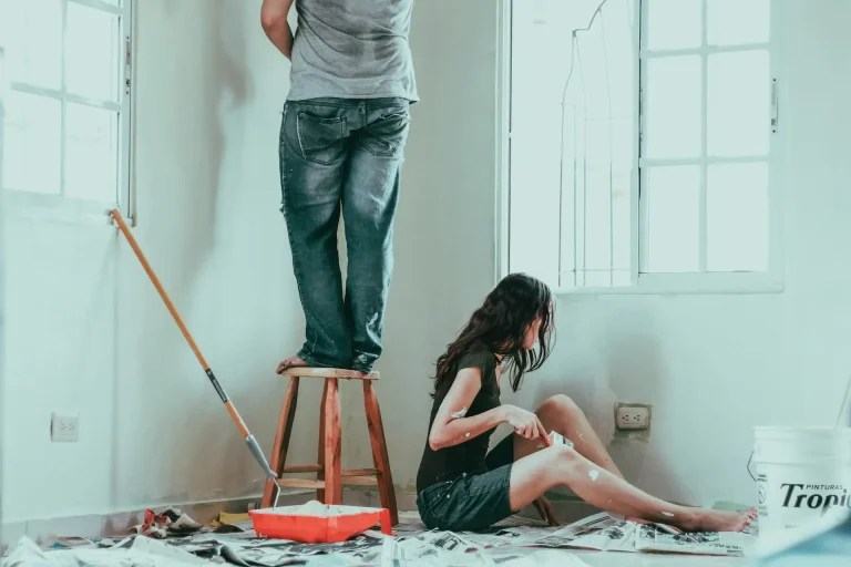 Two people painting a white wall in a bright room with large windows