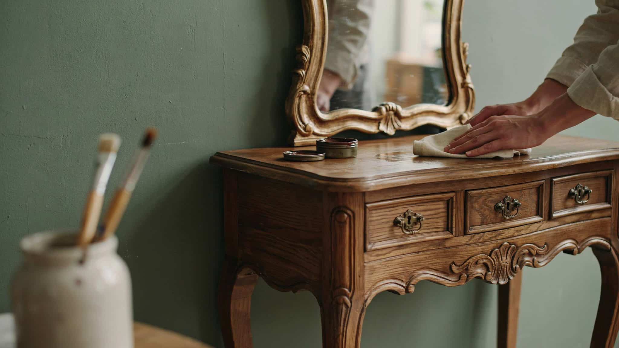 Antique wooden dressing table with ornate mirror in olive green room, being polished by hands during furniture refinishing process in soft natural light.