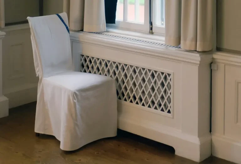 White chair with cover placed near decorative radiator cover under window in softly lit room