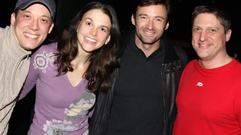 Group photo of Hugh Jackman and Sutton Foster smiling with two other people at an event, standing close together against a dark background.