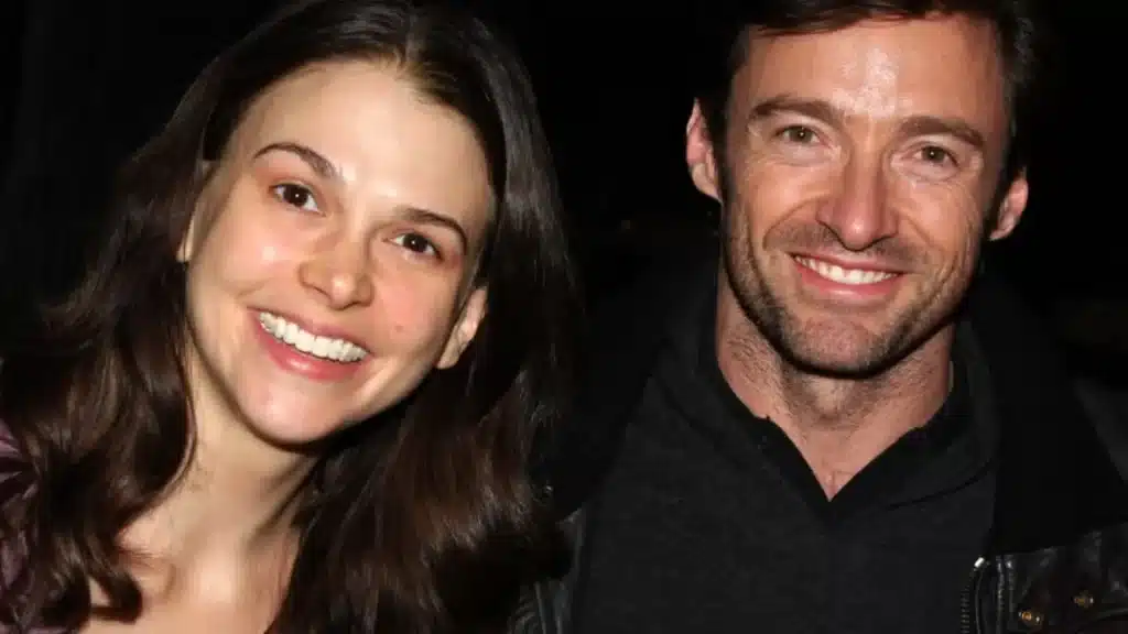 Hugh Jackman and Sutton Foster smiling together in a close-up photo, both looking at the camera against a dark background.