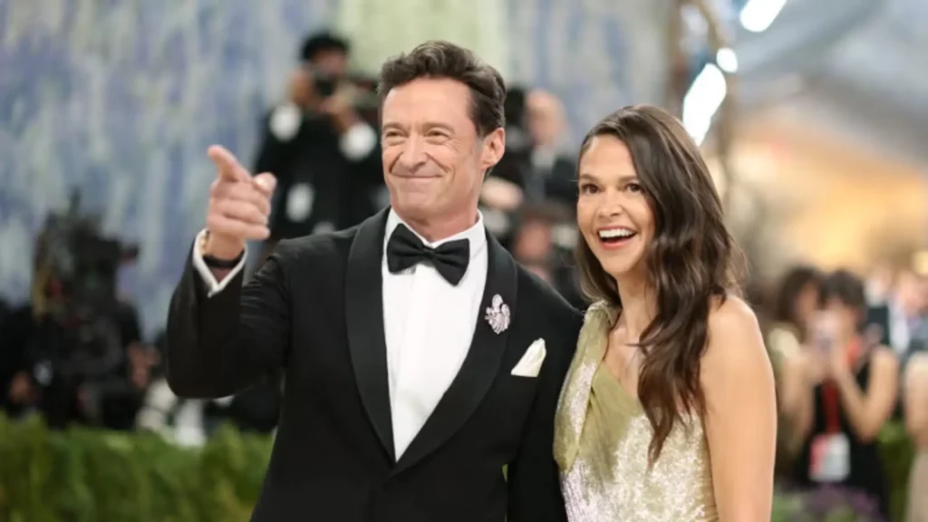 Hugh Jackman and Sutton Foster smiling at a red carpet event, with Hugh gesturing and pointing while Sutton stands beside him laughing, both dressed in formal evening attire.