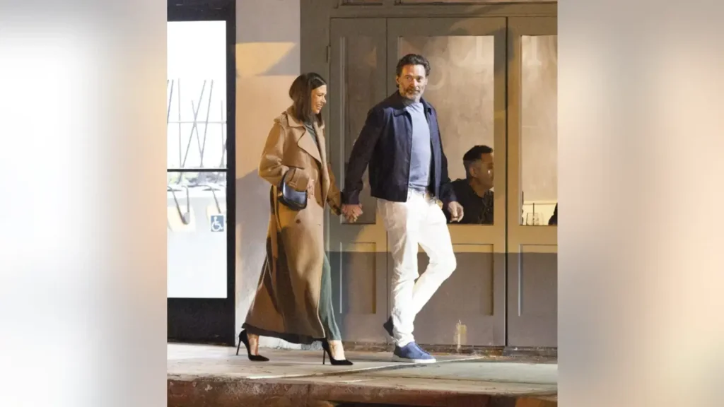 Hugh Jackman and Sutton Foster walking together hand in hand outside a building at night, dressed in casual evening outfits and smiling while leaving a venue.