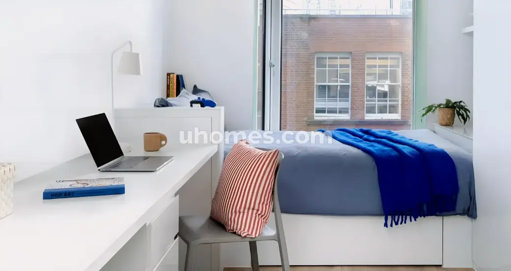 Modern dorm room with white desk, open laptop, gray chair, and bed with blue bedding