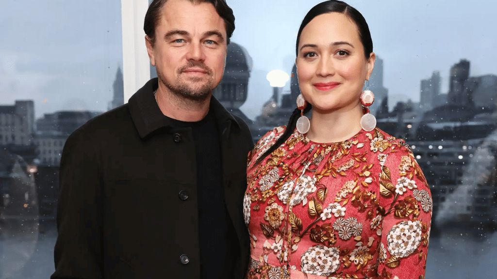 Leonardo DiCaprio and Lily Gladstone standing side by side, posing for a photo at a film event with a city backdrop visible through the window.