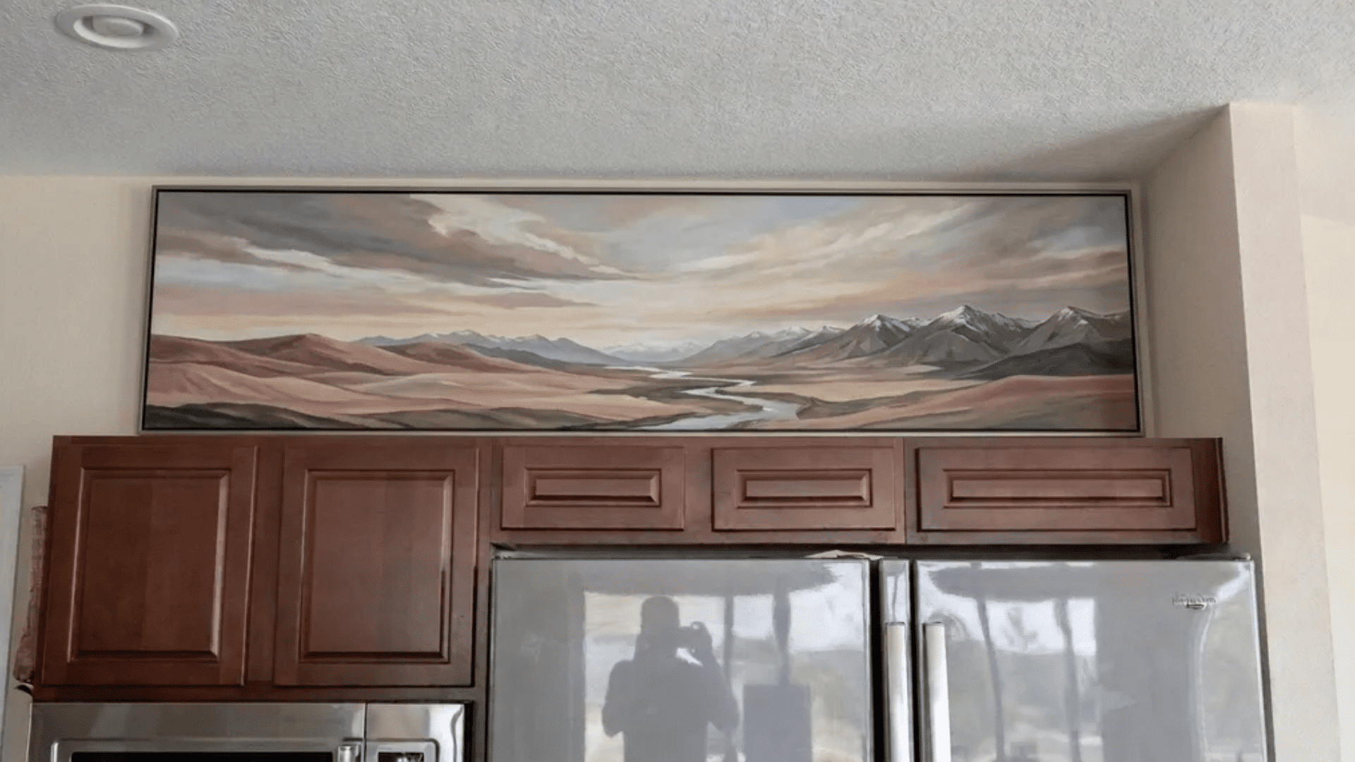 Kitchen wall with a wide landscape painting above wooden cabinets and a stainless steel refrigerator.