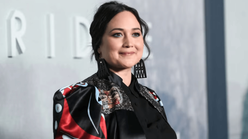 Lily Gladstone smiling at a red carpet event wearing a black outfit with patterned details and large geometric earrings, with a blurred event backdrop behind her