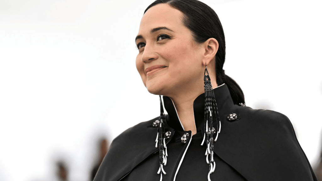 Lily Gladstone in a black outfit with long earrings, smiling in a close-up profile against a light background.