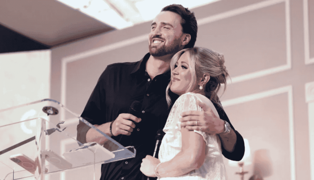 Riley Gaines and her husband embracing on stage at public event holding microphones near podium during emotional celebration moment indoor lighting.