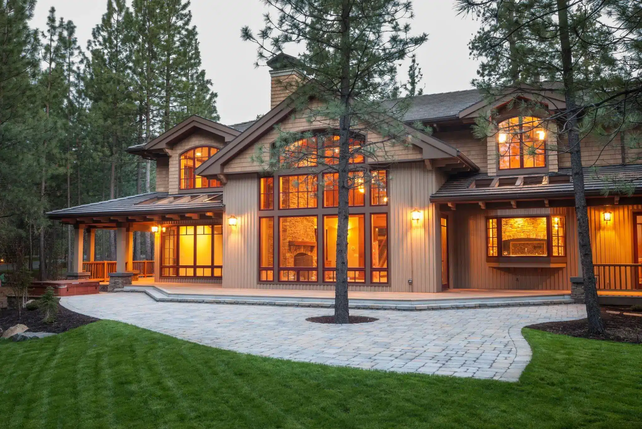Large rustic house with expansive windows illuminated at dusk surrounded by pine trees and stone pathway