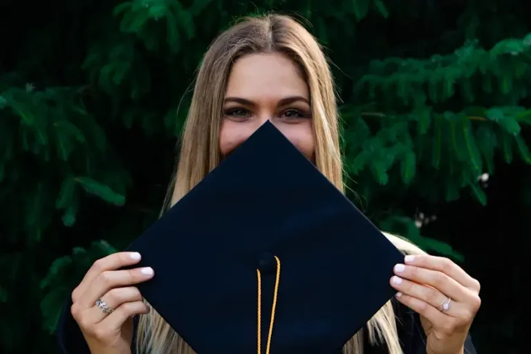 Graduating student holding black cap in front of face outdoors with green tree background