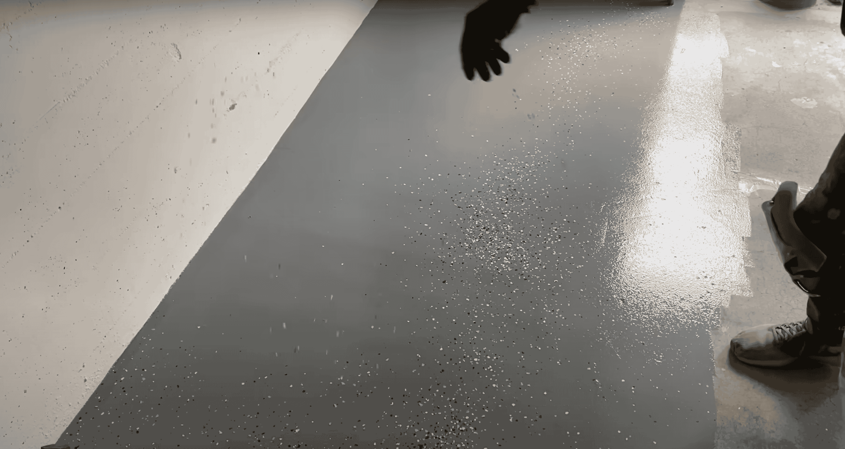 A person scattering dark decorative paint flakes onto a freshly coated grey epoxy garage floor.