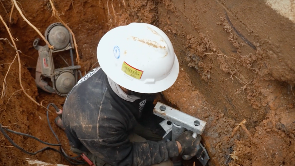 A worker in a hard hat inside an excavation pit, securing a heavy steel pier bracket underneath a concrete foundation footer.