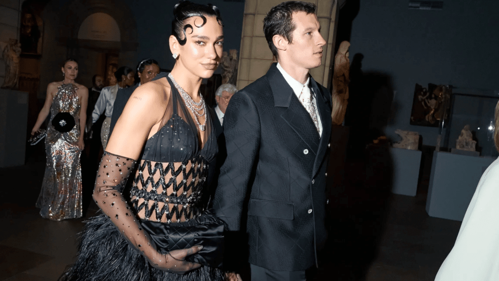 “Dua Lipa and Callum Turner walking at the Met Gala, showcasing her engagement ring and layered jewelry on a black gown