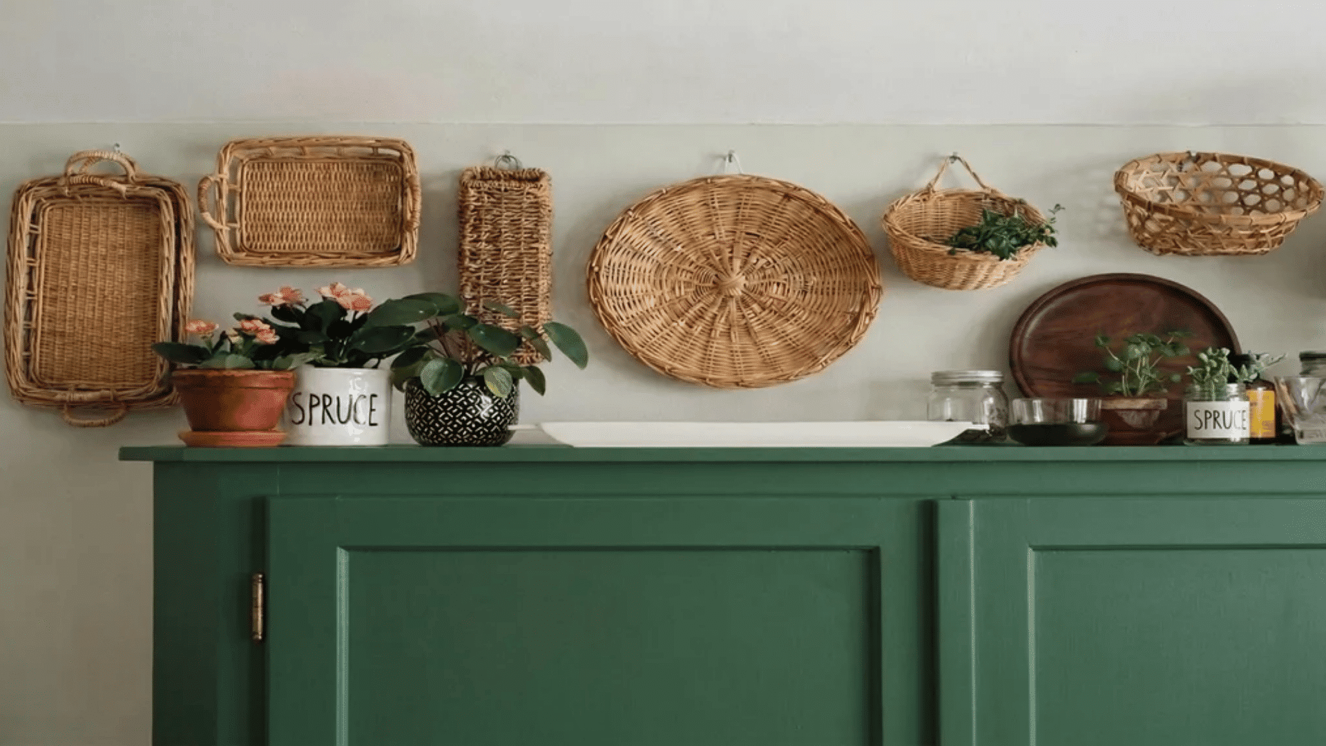 Rattan trays and baskets hung on a wall above a green cabinet with potted plants and jars on top.