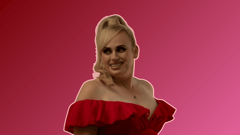 Australian actress Rebel Wilson smiling and posing in a red off-the-shoulder dress against a solid pink background.