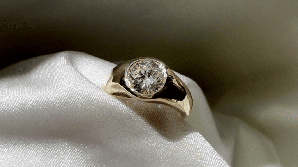 Close-up of a round diamond set low in a wide 18k yellow gold engagement ring on a white satin surface