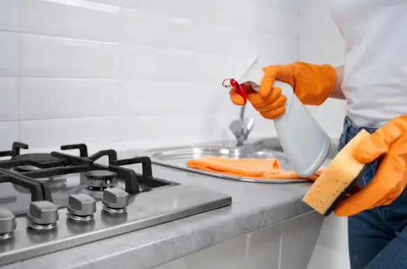 Person cleaning kitchen countertop with spray bottle and sponge wearing orange gloves