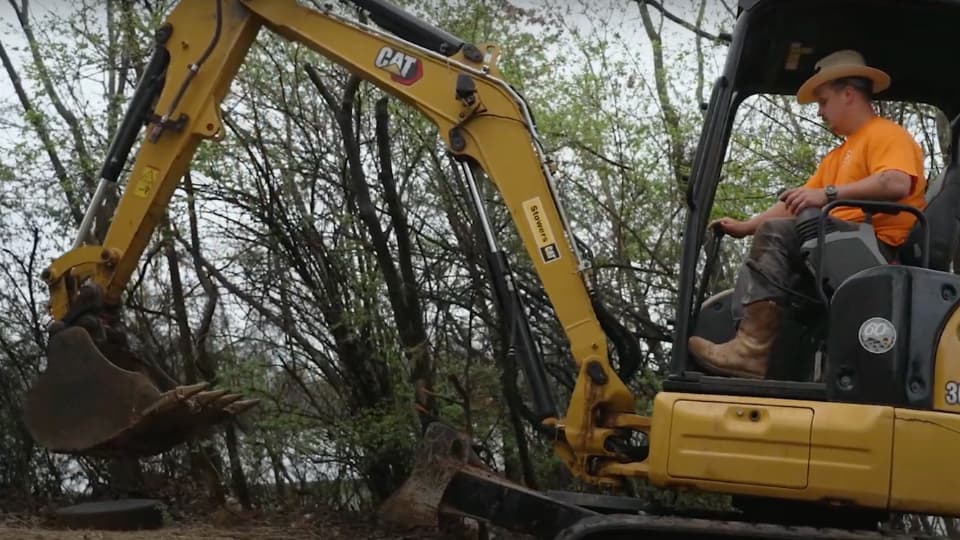 A worker operating a yellow mini excavator to clear soil and debris near trees on a construction site.