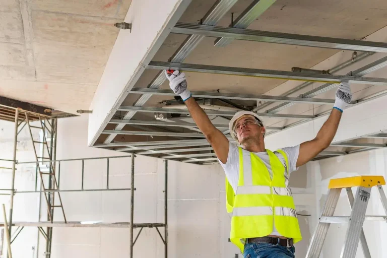 Construction worker in high-visibility vest installing ceiling framework indoors with ladder and scaffolding nearby