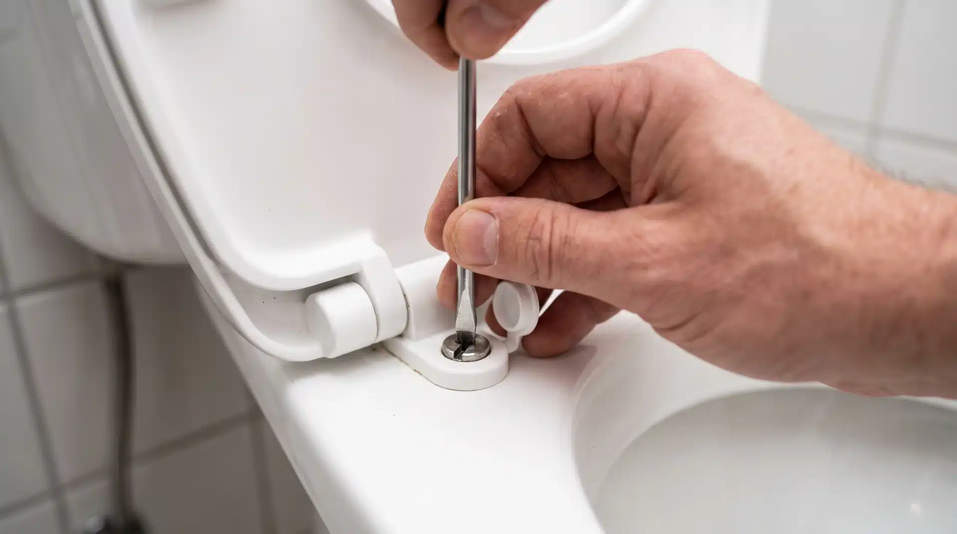 Hand using a flathead screwdriver to unscrew a mounting bolt on a white toilet seat hinge.