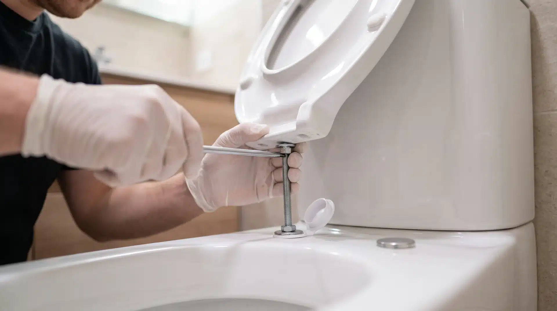 Hand tightening a mounting bolt on a newly installed white toilet seat with a flathead screwdriver.