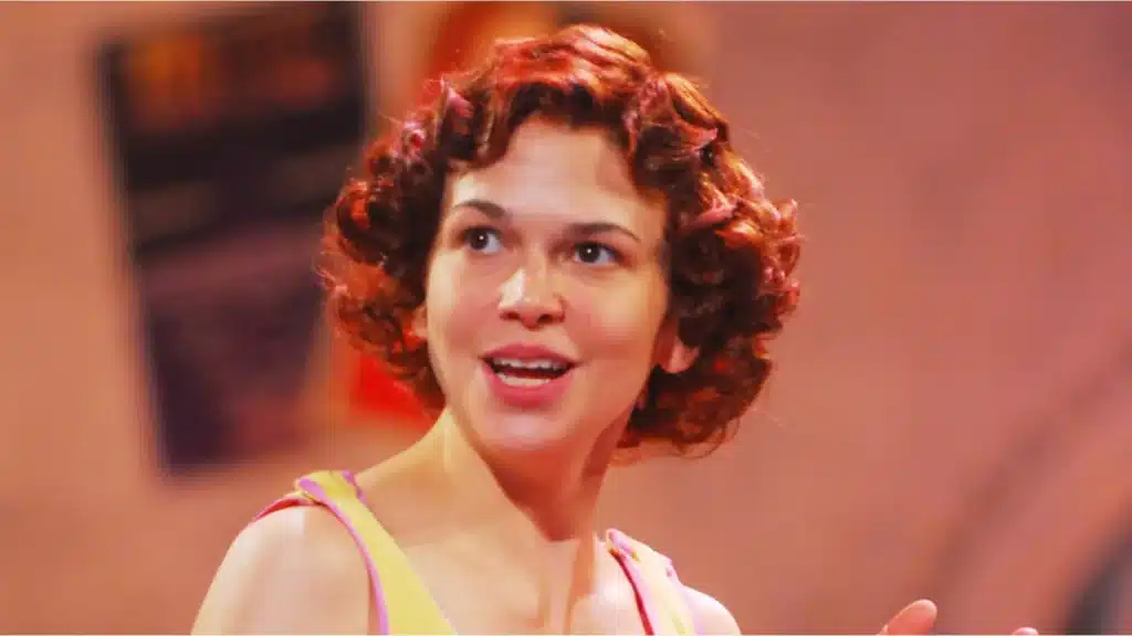Sutton Foster on stage, smiling and looking off to the side while wearing a colorful performance costume with curled short hair under warm theatrical lighting.