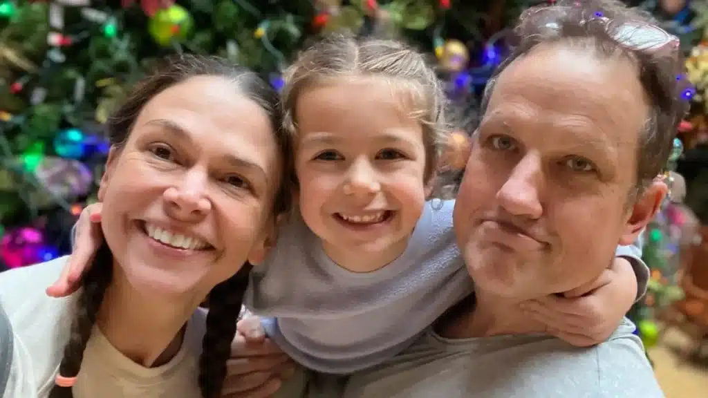 Sutton Foster and Ted Griffin smiling with a young child in front of a decorated Christmas tree, in a warm family-style holiday photo.