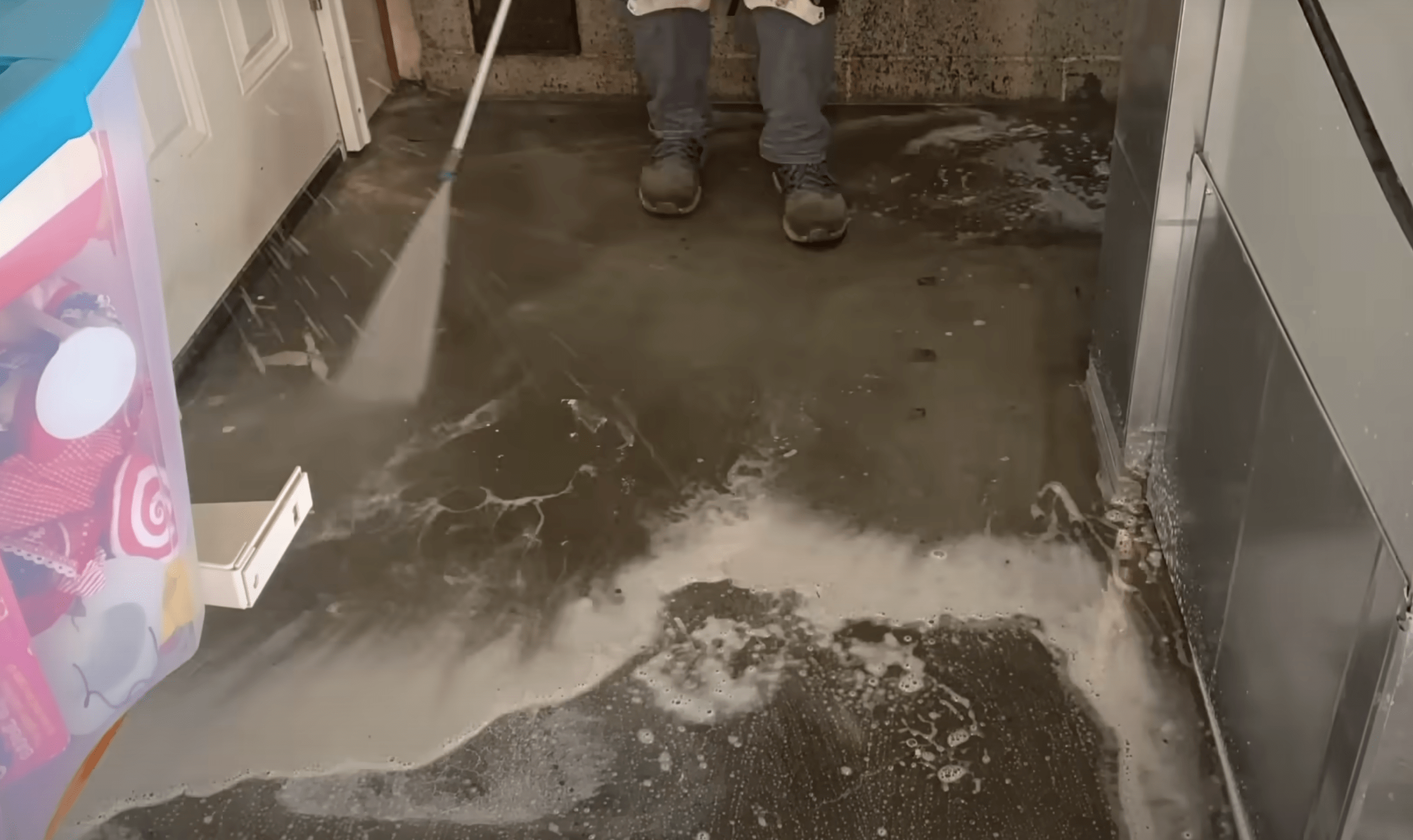 A person power washing a stained garage concrete floor, spraying down soapy water and debris.