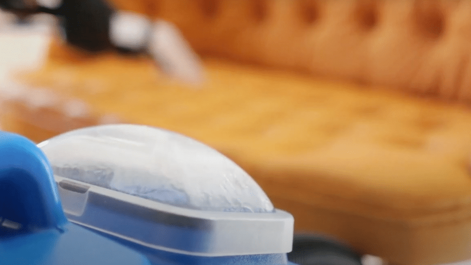 A person applying a cleaning solution from a blue container onto a small patch of tan outdoor carpet to test it.