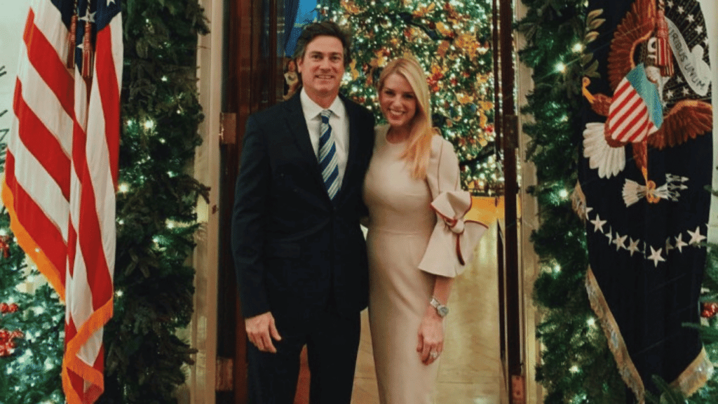 Two people standing together in formal clothing near Christmas decorations and American flags inside a building.