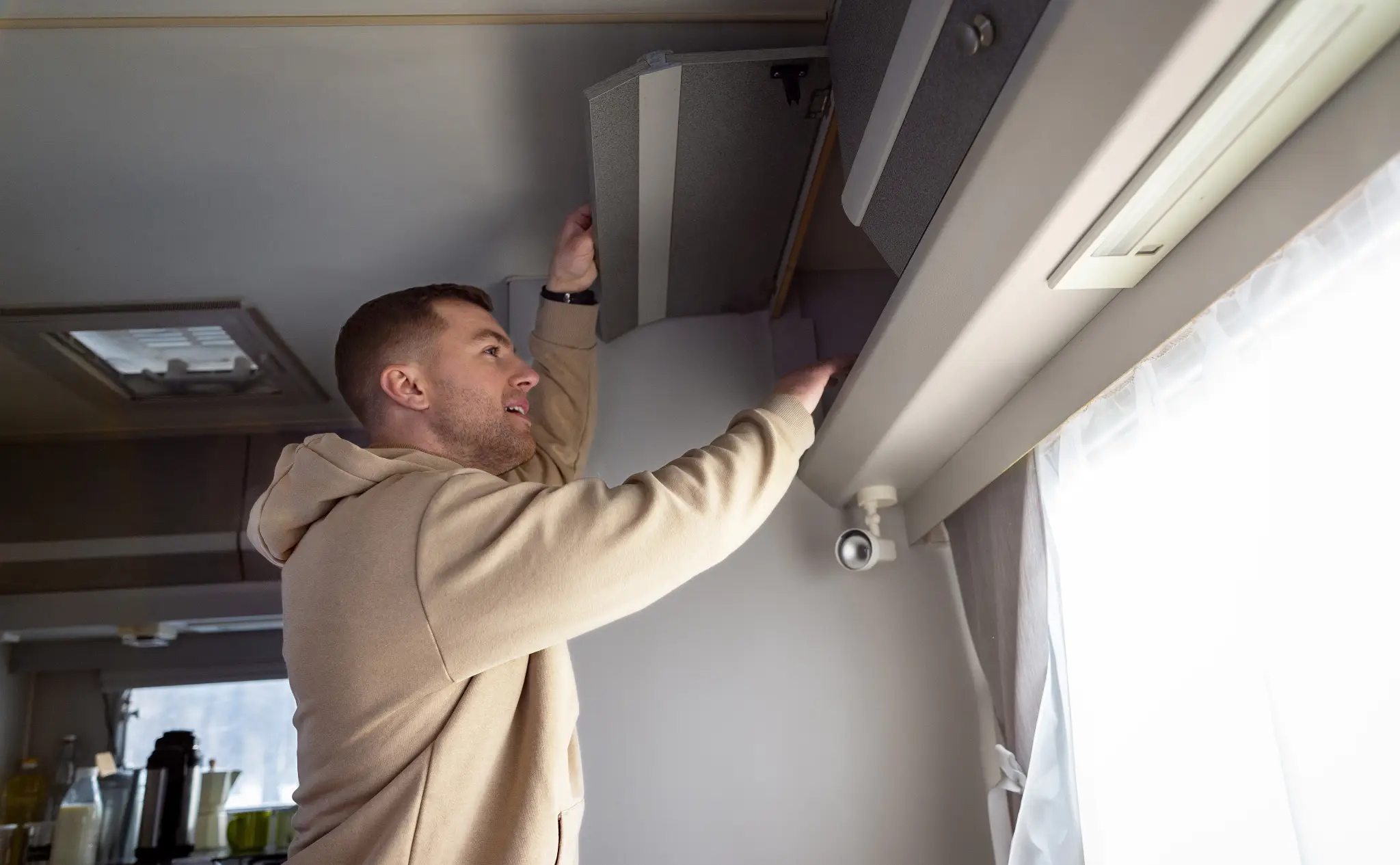 Man reaching into overhead cabinet in RV interior, wearing beige hoodie, bright daylight through window