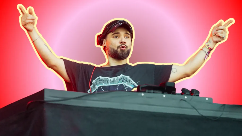 Skrillex performing behind DJ decks with arms raised at a live festival show.