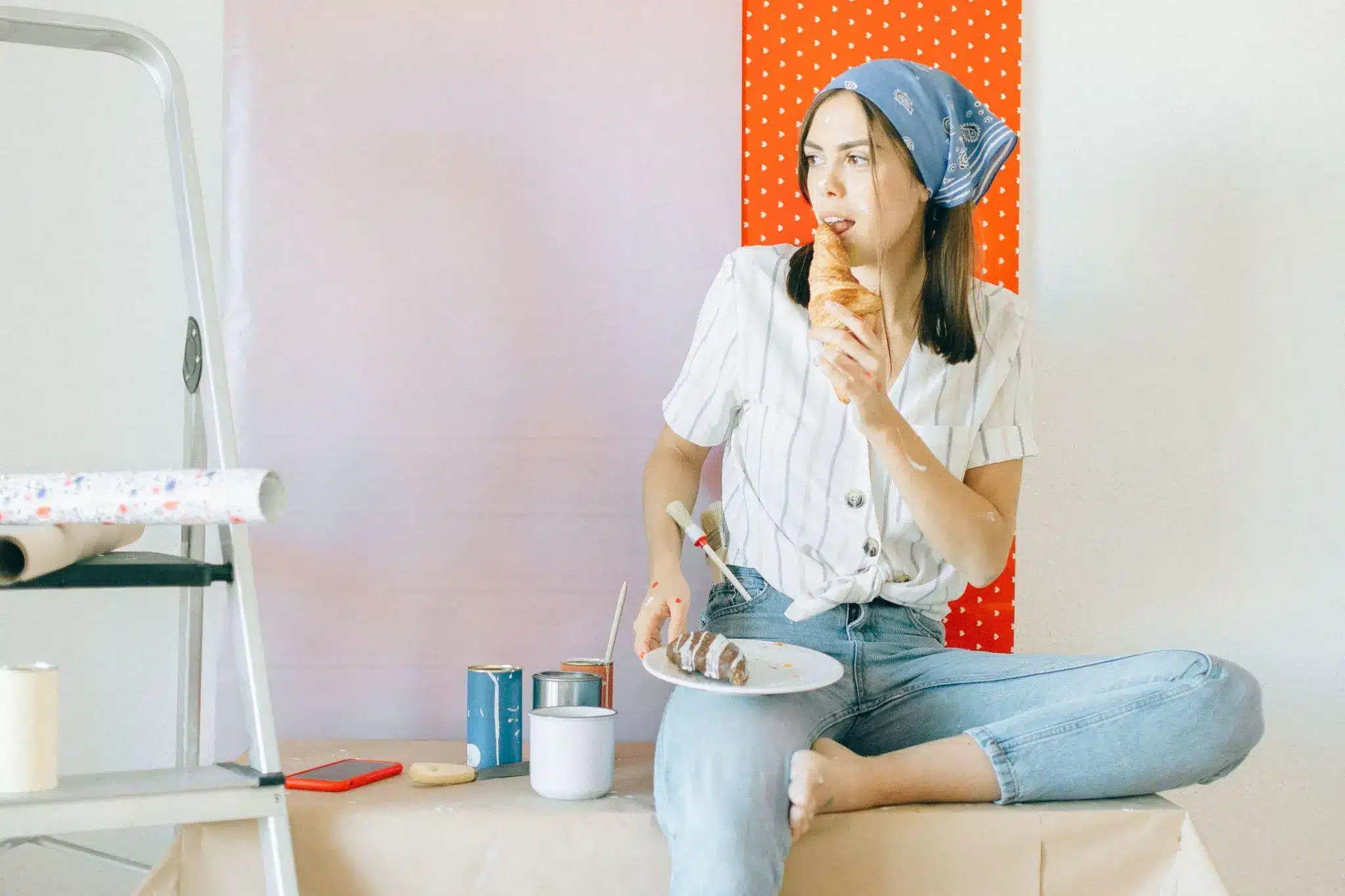 Woman sitting on table eating croissant with paint cans and brushes nearby in casual setting