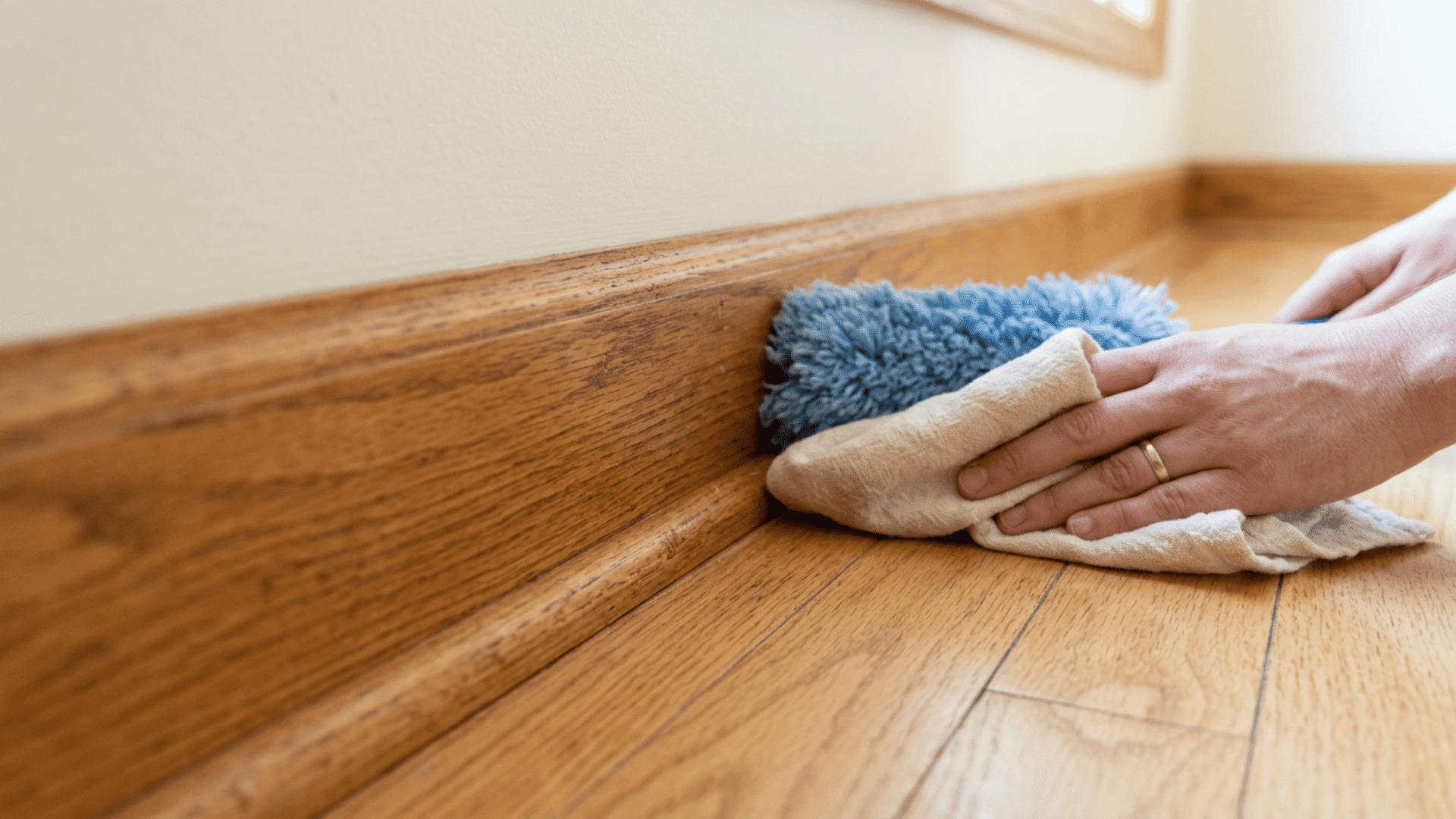 Wood baseboard being dusted and wiped with a damp cloth near floor edge