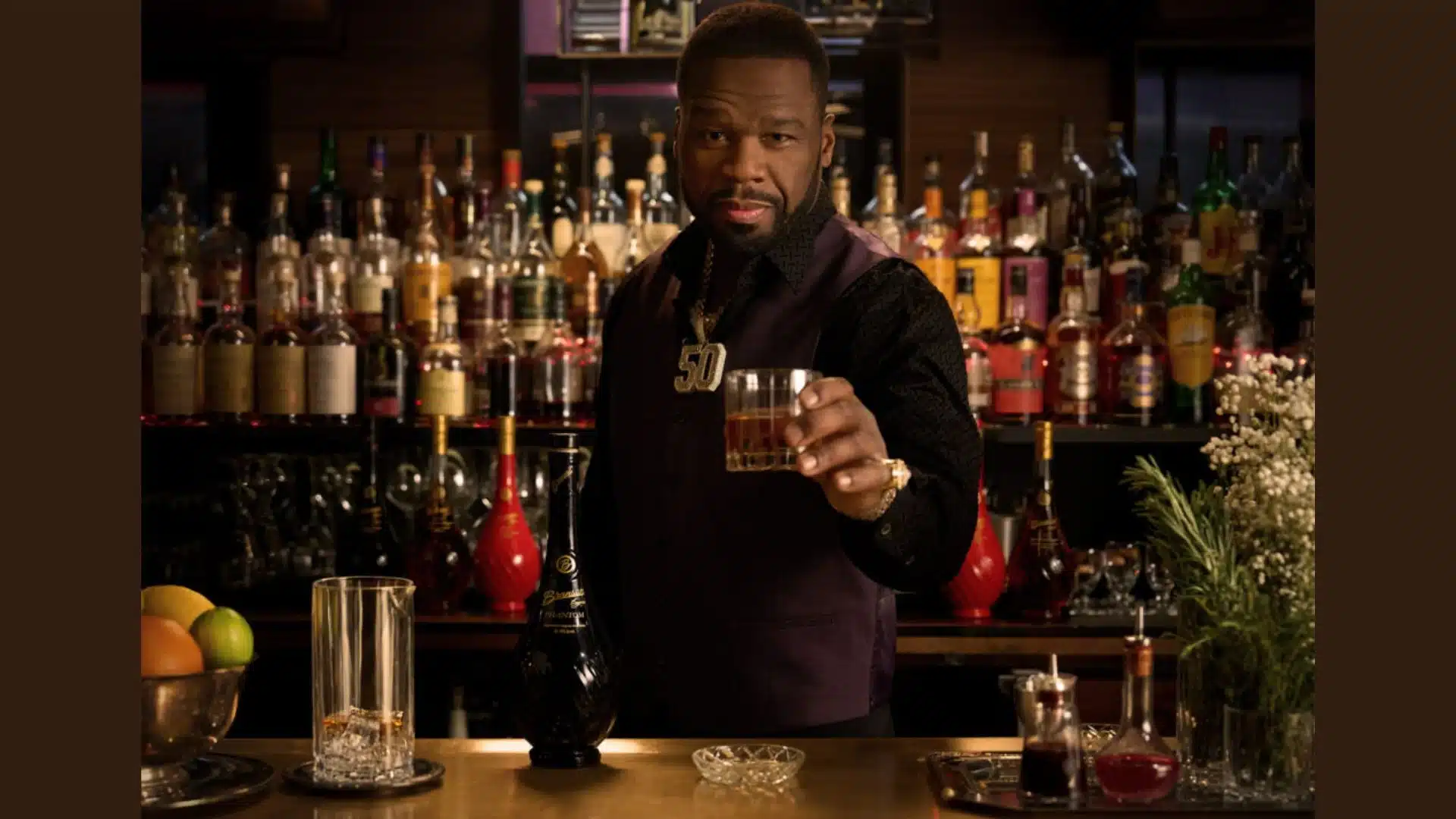 50 Cent holding a glass of liquor at a bar surrounded by spirits bottles