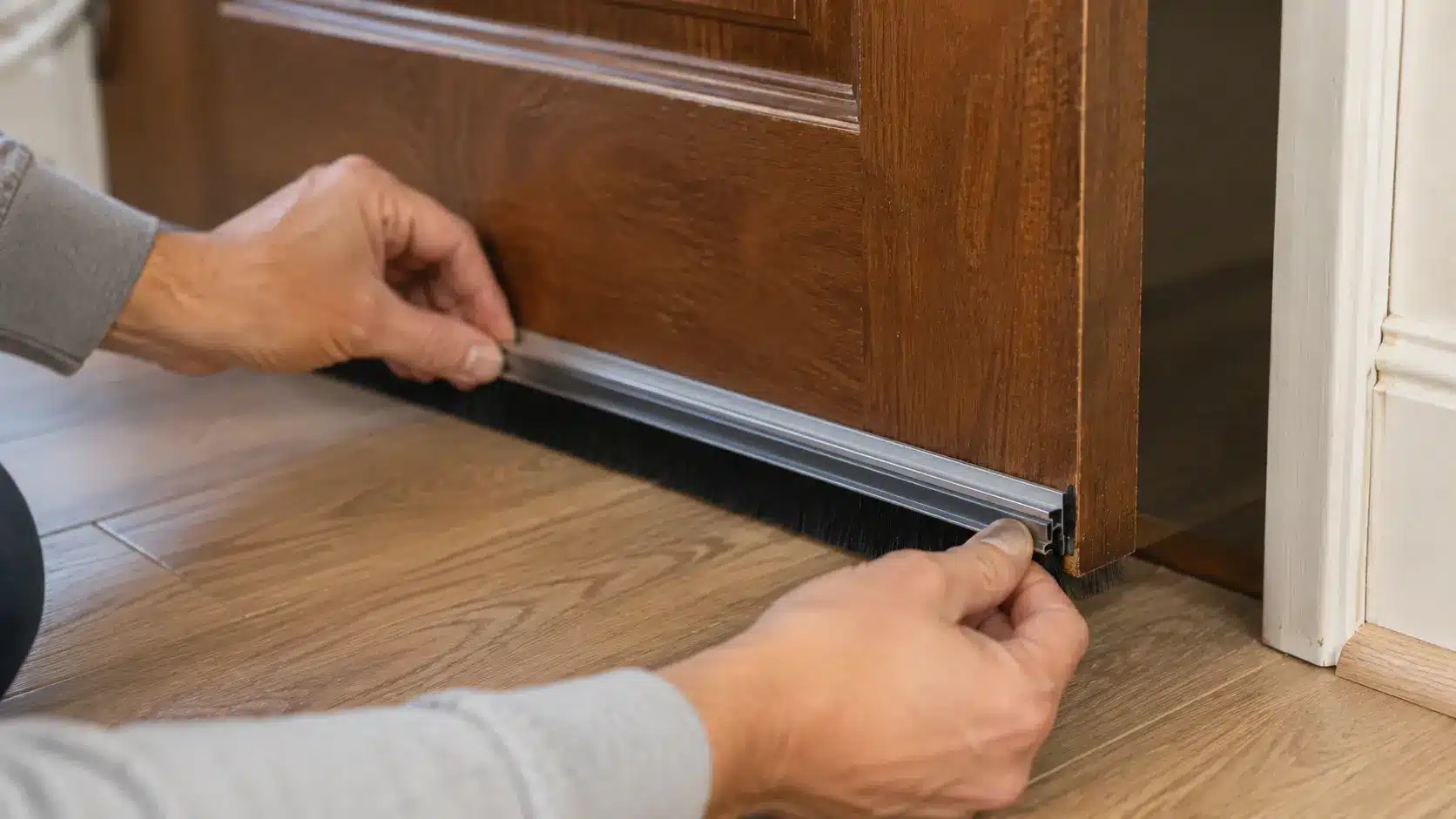 Person installing a brush door sweep along the bottom of a wooden door to block sound and drafts.
