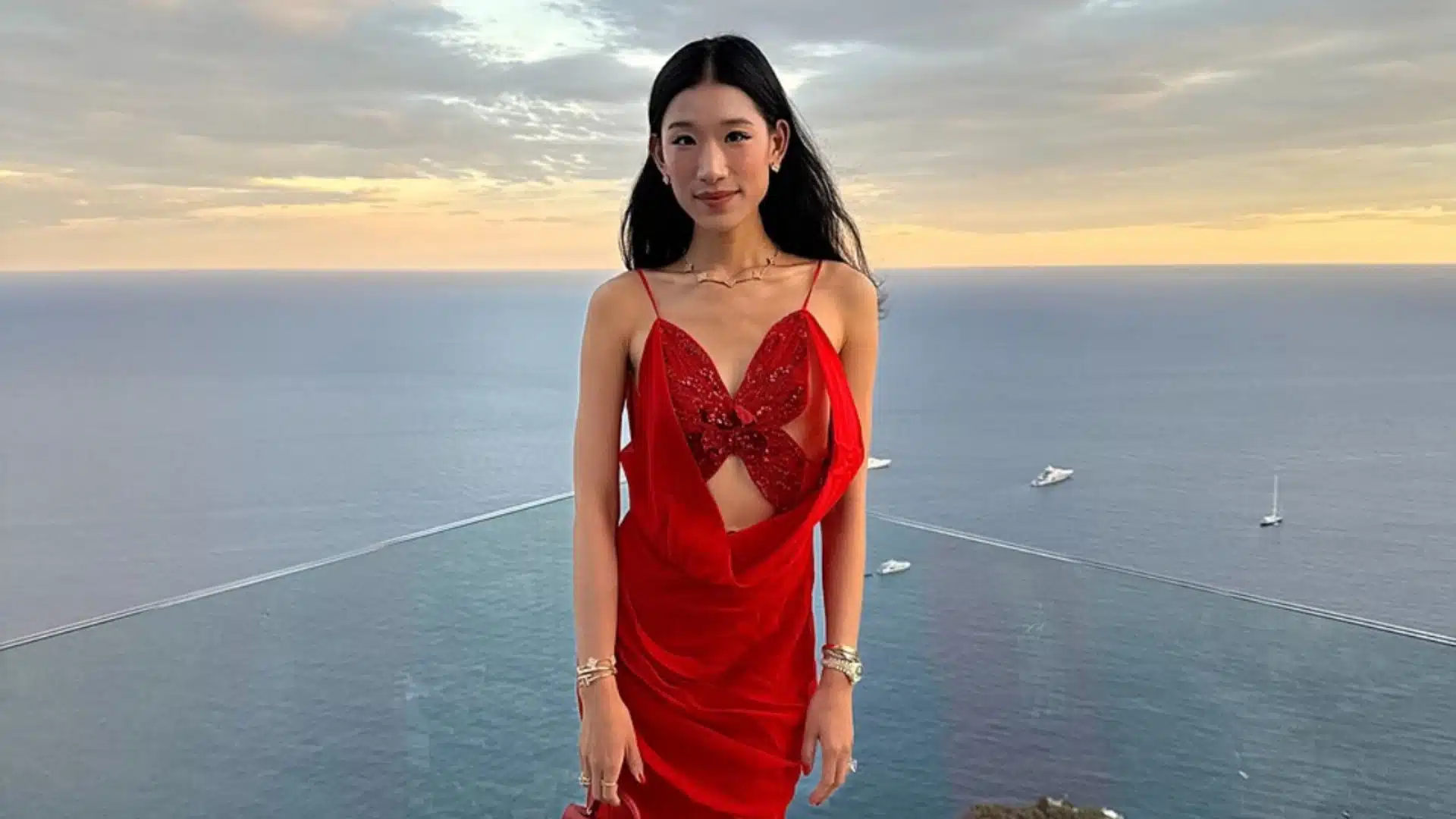 Becca Bloom wearing a red dress on a seaside terrace at sunset.