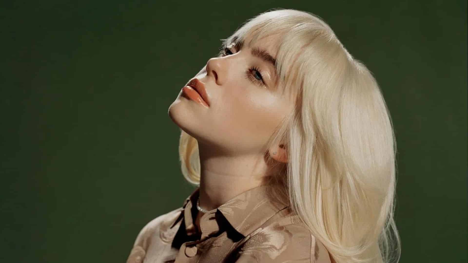 Billie Eilish with long blonde hair posing against a dark green background